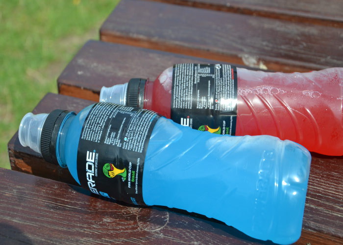 red and blue bottles of sports drinks with sugar lay on a brown bench outside by the grass